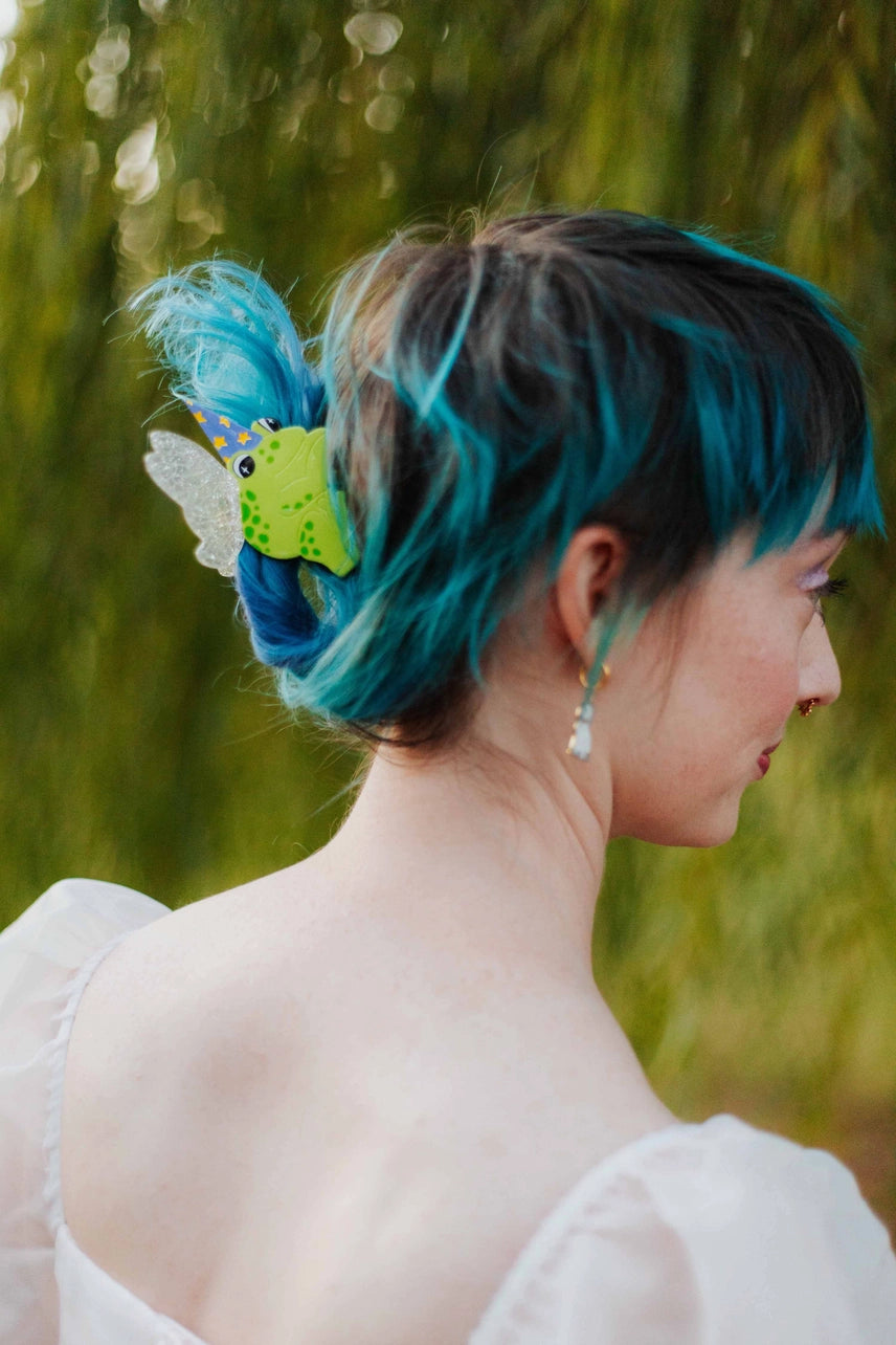 Person with teal hair styled in an updo with a decorative hairpiece against a blurred green background
