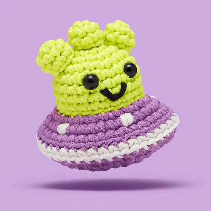 Crocheted alien with a green face and purple spaceship on a purple background