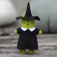 A felt cat toy dressed in a witch costume, featuring a green body, a black dress with a white collar, and a black witch hat, set against a gray wooden background.