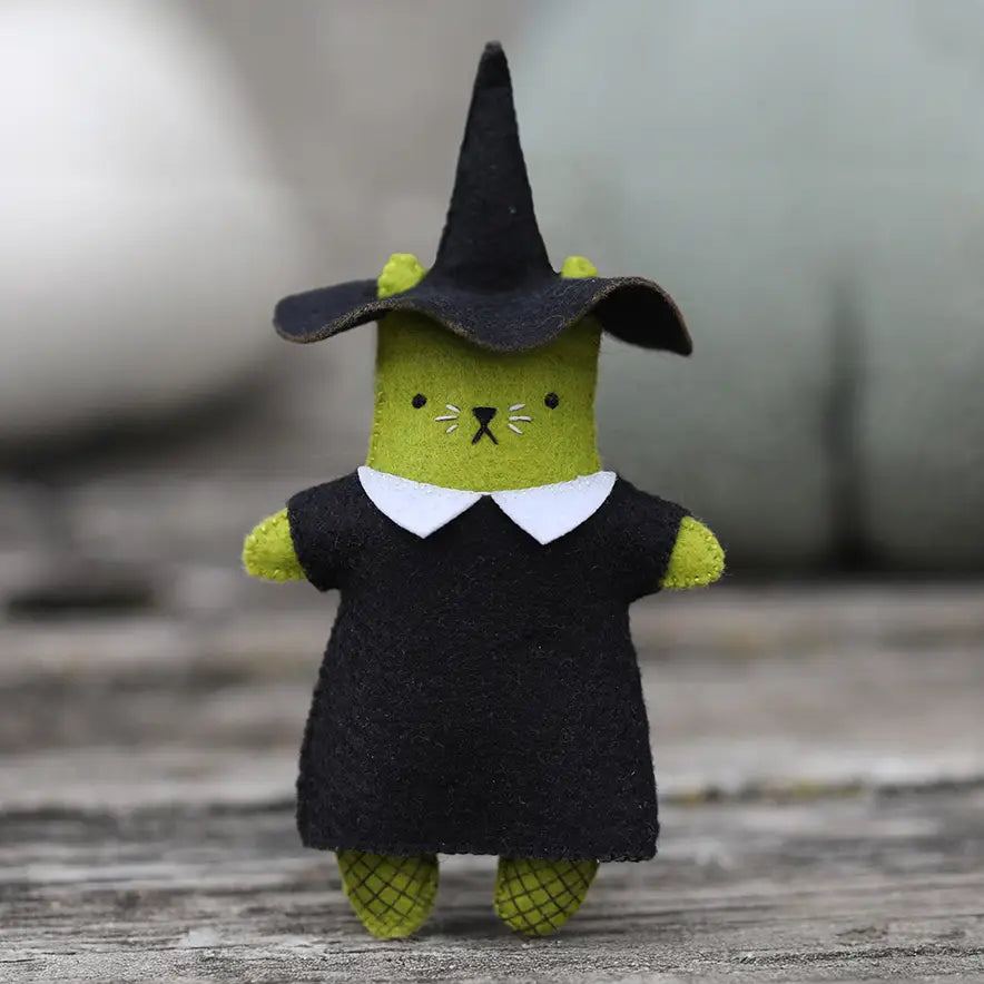 A felt cat toy dressed in a witch costume, featuring a green body, a black dress with a white collar, and a black witch hat, set against a gray wooden background.