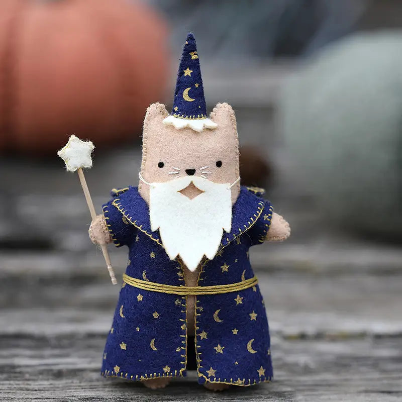 A felt cat toy dressed in a wizard costume, featuring a beige body, a blue robe and hat with gold moons and starts, and a wand with a star on top, set against a gray wooden background.