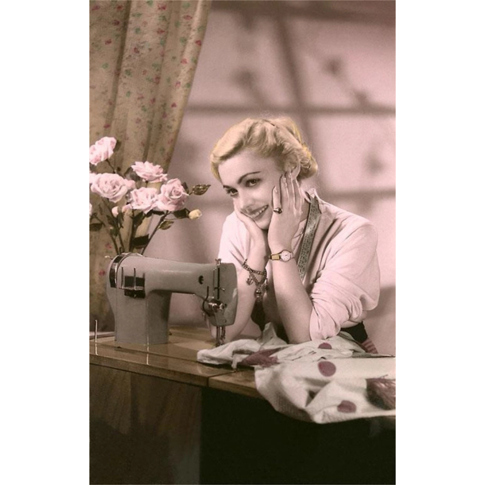 Woman smiling and staring off into space with hands to her facing while seated at a table with roses, sewing machine, and blanket. The background has a floral curtain hanging and the shadows of window panes