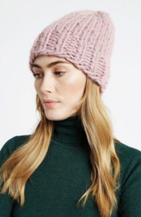 Model with blonde hair and green turtleneck wearing a light mauve knit hat on a white background.