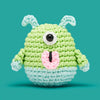 Crocheted green and blue creature with a single eye on a teal background