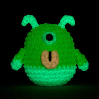 Green crocheted alien with a single eye glowing in the dark on a black background